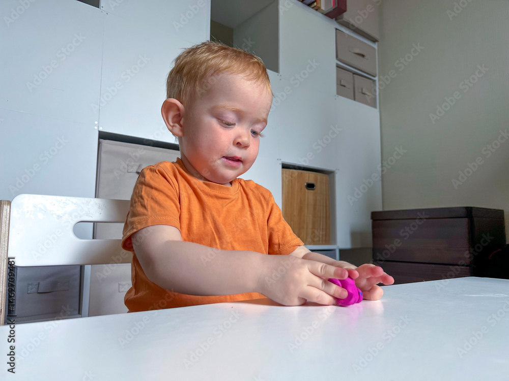 A red-haired boy in an orange T-shirt sculpts from colored plasticine at a table in a room. Development of fine motor skills according to the Montessori system