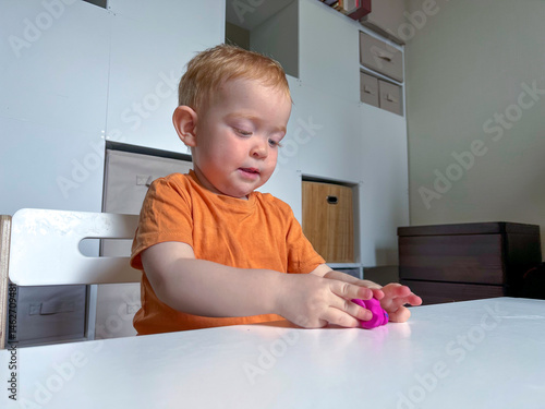 Obraz na płótnie A red-haired boy in an orange T-shirt sculpts from colored plasticine at a table in a room. Development of fine motor skills according to the Montessori system w salonie