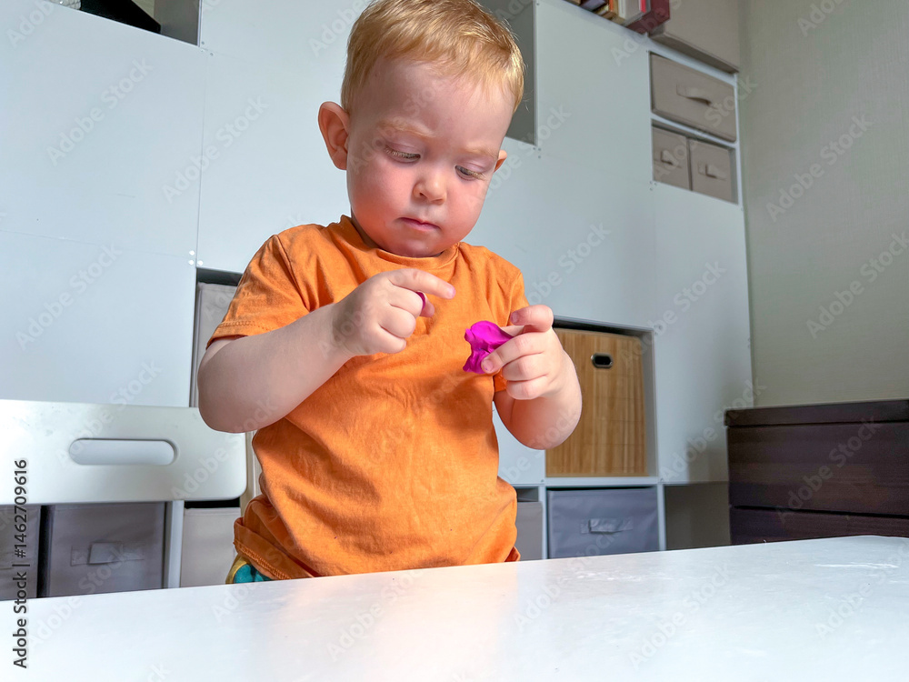 A red-haired boy in an orange T-shirt stands in front of a table and sculpts from plasticine. Early development of a child at home