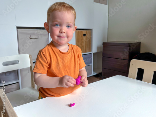 Obraz na płótnie A red-haired boy in an orange T-shirt stands in front of a table and sculpts from plasticine. Early development of a child at home w salonie