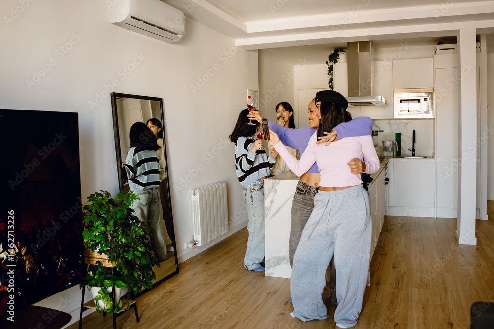Fototapeta premium Two female friends standing and smiling while posing in front of a mirror while one of them holds a phone and the other a glass
