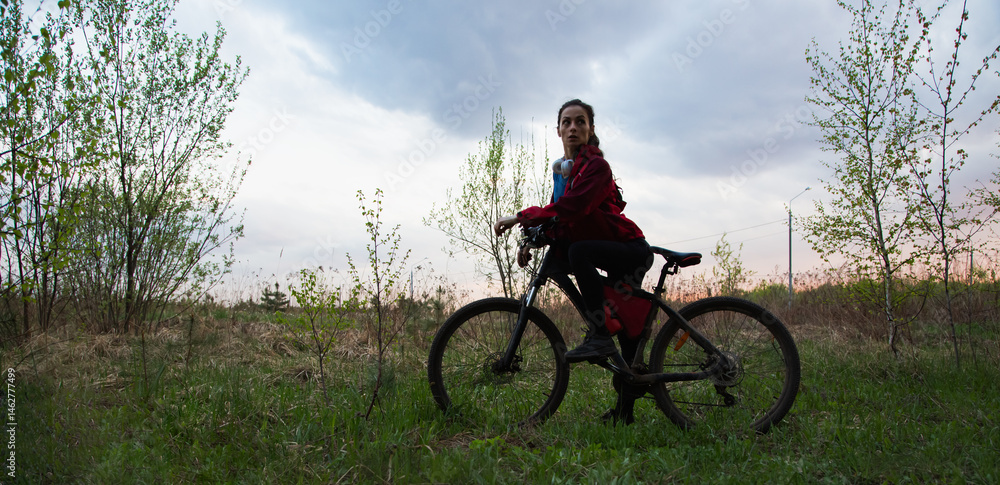 Fototapeta premium Young woman riding a bicycle on the spring field with green grass on sunset sky landscape background. Happy activity sport trip vacations