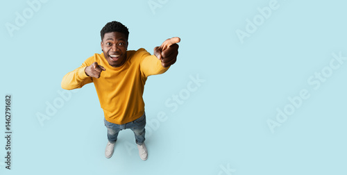 Happy african american young man in casual showing copy space, pointing at blank space for advertisement or text, blue studio background, panorama, top view, full length shot