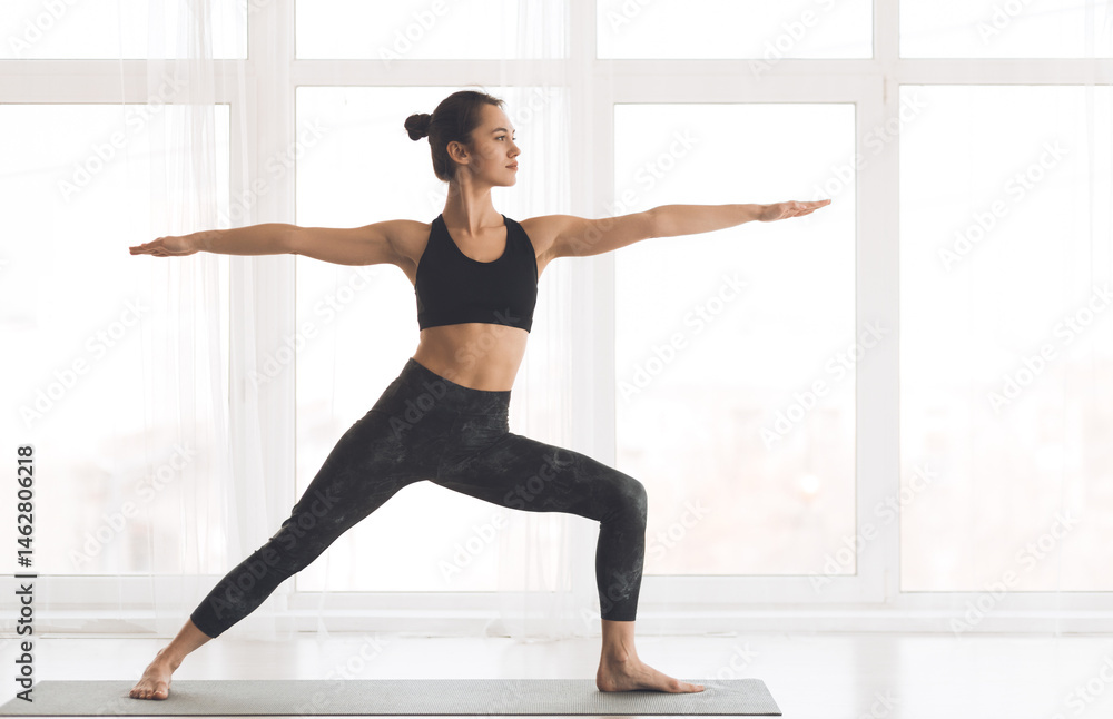 Naklejka premium Virabhadrasana Pose. Fit girl standing by window in warrior pose in modern studio, copyspace, full length
