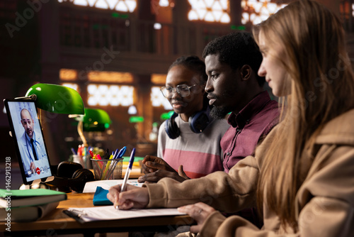 Diverse academic scholars in a library join a video call with a medic tutor, enhancing their knowledge of science, treatment methods and telemedicine. Students learn about medical practice lessons.