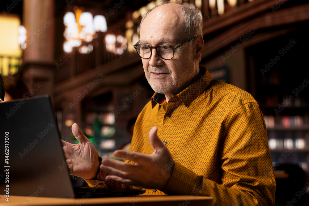 Senior man involved in a remote classroom experience via online platform, using wireless internet to study a university course and chatting with his teacher. Reviewing project materials.