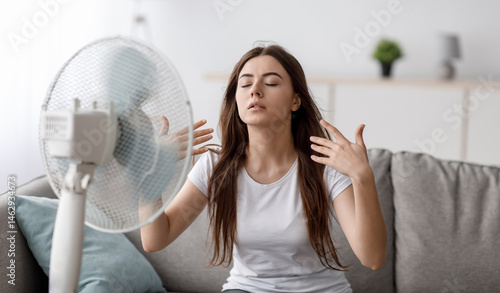 Canvastavla Sad young european woman suffering from heat sits on sofa, catches cold air from fan and waves hands to herself in living room interior