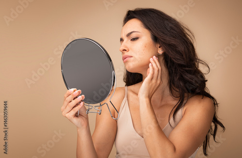 Obraz na płótnie Stressed sad brunette young woman wearing beige top looking at mirror and touching her face over beige studio background, having skin problems