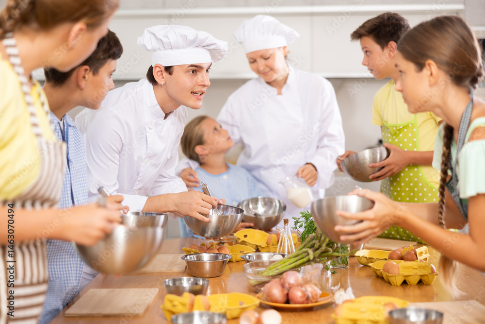 Enthusiastic young chef giving culinary classes to group of interested curious tween girls and boys, teaching to cook deliciously..