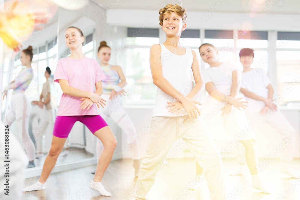 Fototapeta premium Male teen performs choreographic exercises and teaches energetic mobile social house dance together with friends. Young girls and guy in repeat movements, train in spacious studio