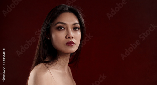 Studio portrait of a beautiful young Asian woman with long dark hair and bare shoulders against a dark red background