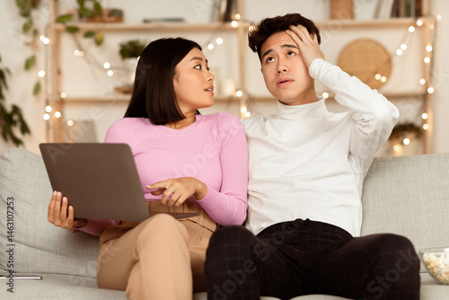 Принти на полотні Asian Shopaholic Wife Shopping Online With Shocked Husband, Asking Him Buy Expensive Clothes And Spend Too Much Money Sitting On Sofa At Home