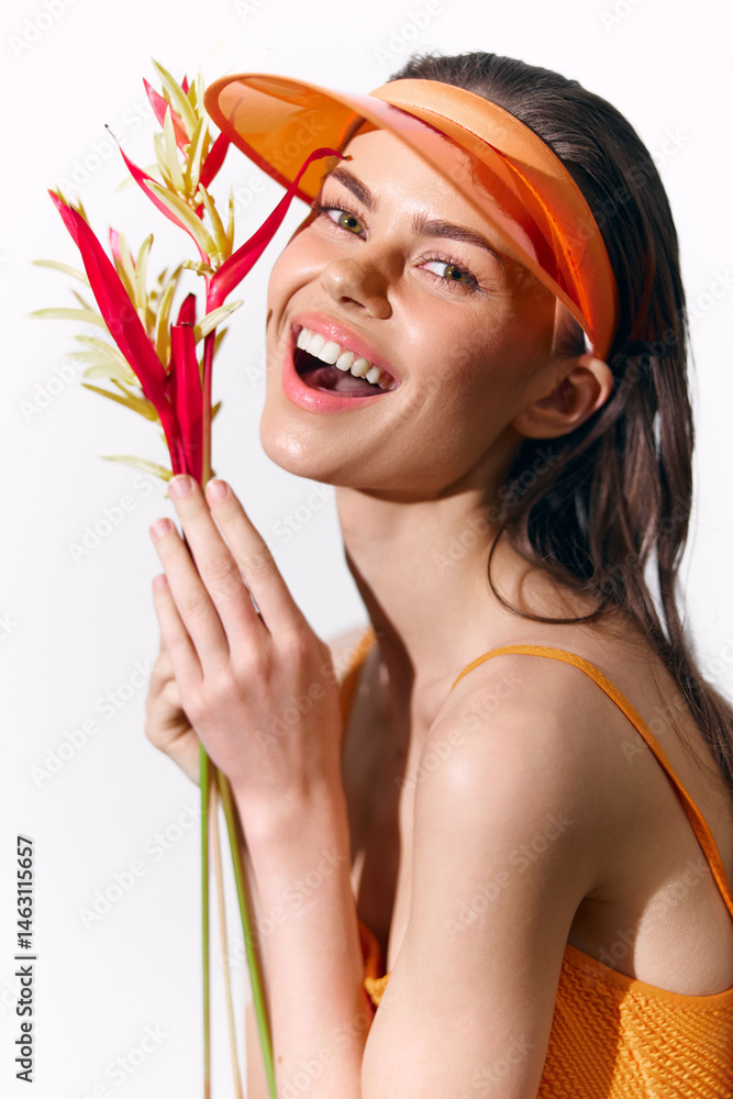Cheerful young woman holding tropical flowers in vibrant orange attire, wearing a sun visor for a summery, joyful vibe.
