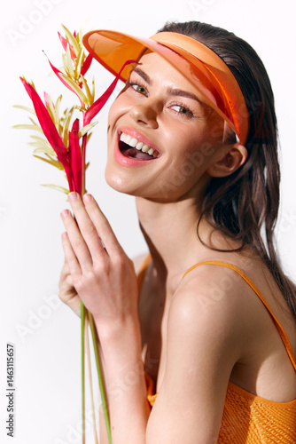 Obraz na płótnie Cheerful young woman holding tropical flowers in vibrant orange attire, wearing a sun visor for a summery, joyful vibe. w salonie