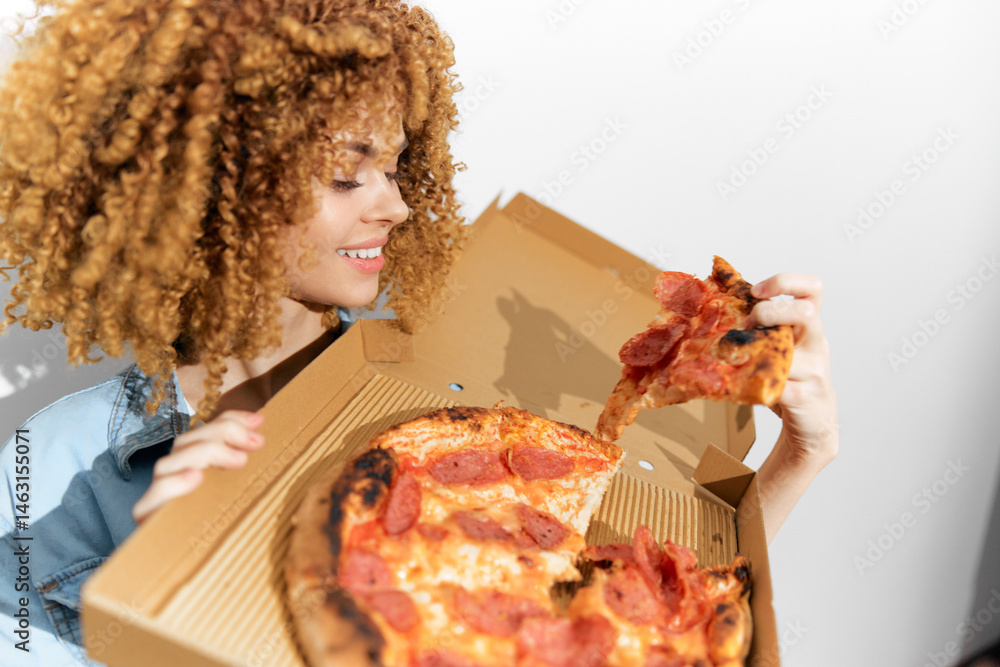 Happy woman with curly hair enjoying delicious pepperoni pizza, holding a slice from a takeout box, showcasing vibrant colors and joyful dining experience