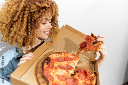 Obraz na płótnie Happy woman with curly hair enjoying delicious pepperoni pizza, holding a slice from a takeout box, showcasing vibrant colors and joyful dining experience w salonie