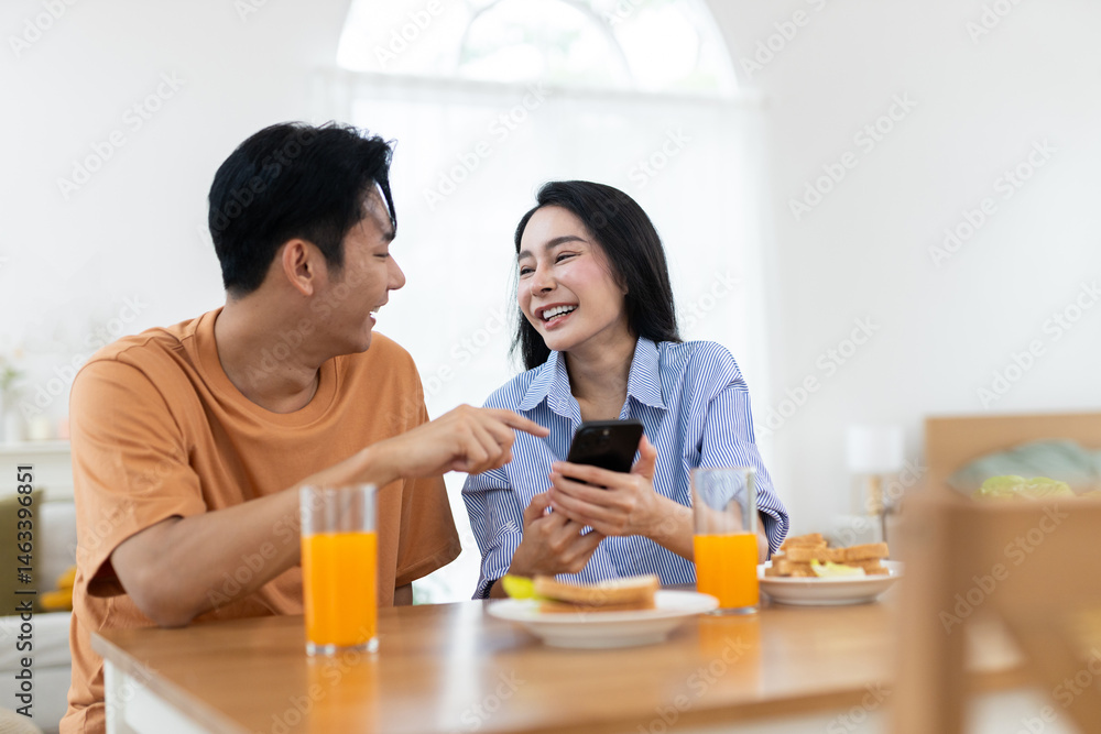 Couple enjoying together while laughing look at smartphone in dining table with glasses of orange juice and breakfast items are present in bright bedroom background. Couple enjoying together while laughing look at smartphone in dining table with glasses of orange juice and breakfast items are present in bright bedroom background.