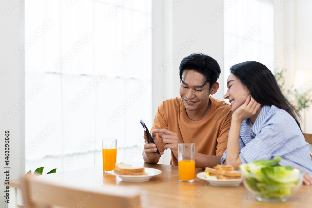 Couple enjoying together while laughing look at smartphone in dining table with glasses of orange juice and breakfast items are present in bright bedroom background. Couple enjoying together while laughing look at smartphone in dining table with glasses of orange juice and breakfast items are present in bright bedroom background.