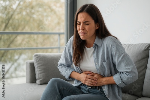 A woman shows a pained expression while clutching her stomach, sitting on a sofa in a striped shirt and jeans