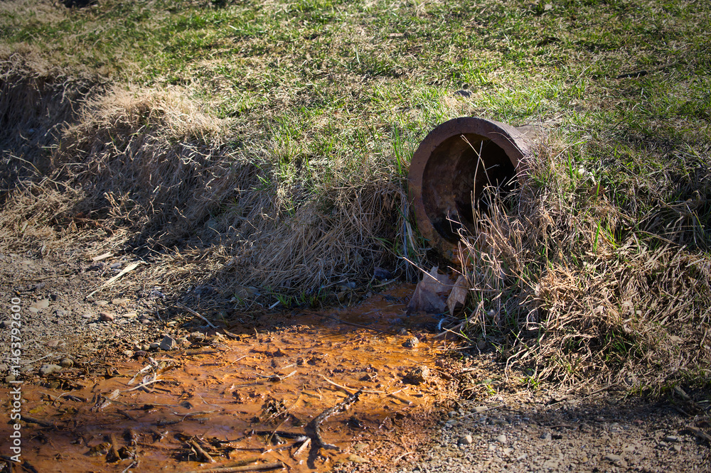 Obraz premium A rusted pipe spills rusty water into a muddy area with weeds and grass growing around it.