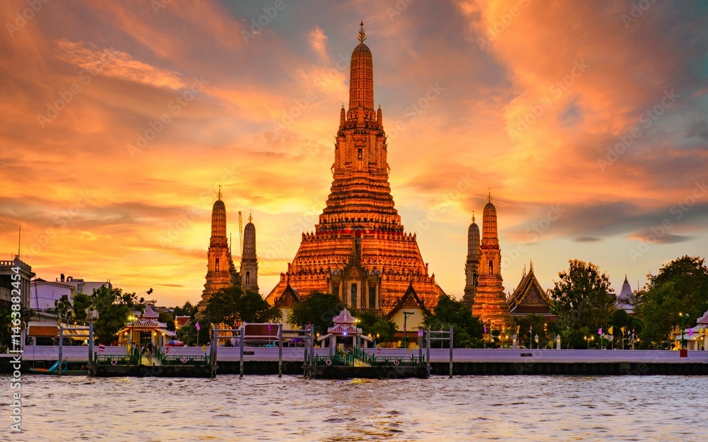 Fototapeta premium Wat Arun stands majestically illuminated by golden rays as twilight envelops Bangkok. Visitors enjoy the stunning architectural details reflecting off the tranquil waters of the Chao Phraya River.