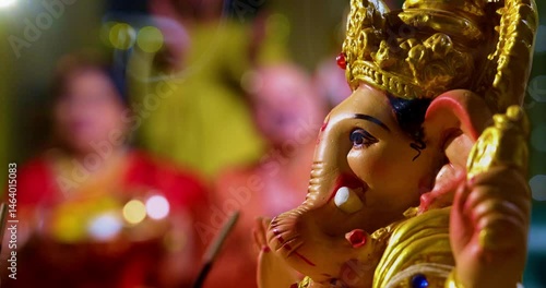 Focus on Lord Ganesha idol during Ganesh Chaturthi as Indian joint family with senior parents, son, daughter-in-law offer prayers with laddu plate and puja thali indoors in decorated festive home