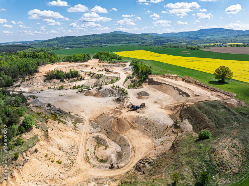 Aerial drone view of sand or gravel quarry surrounded by forest and farmland with distant hills. Aerial Drone view shows an inactive or abandoned quarry embedded in a green landscape.