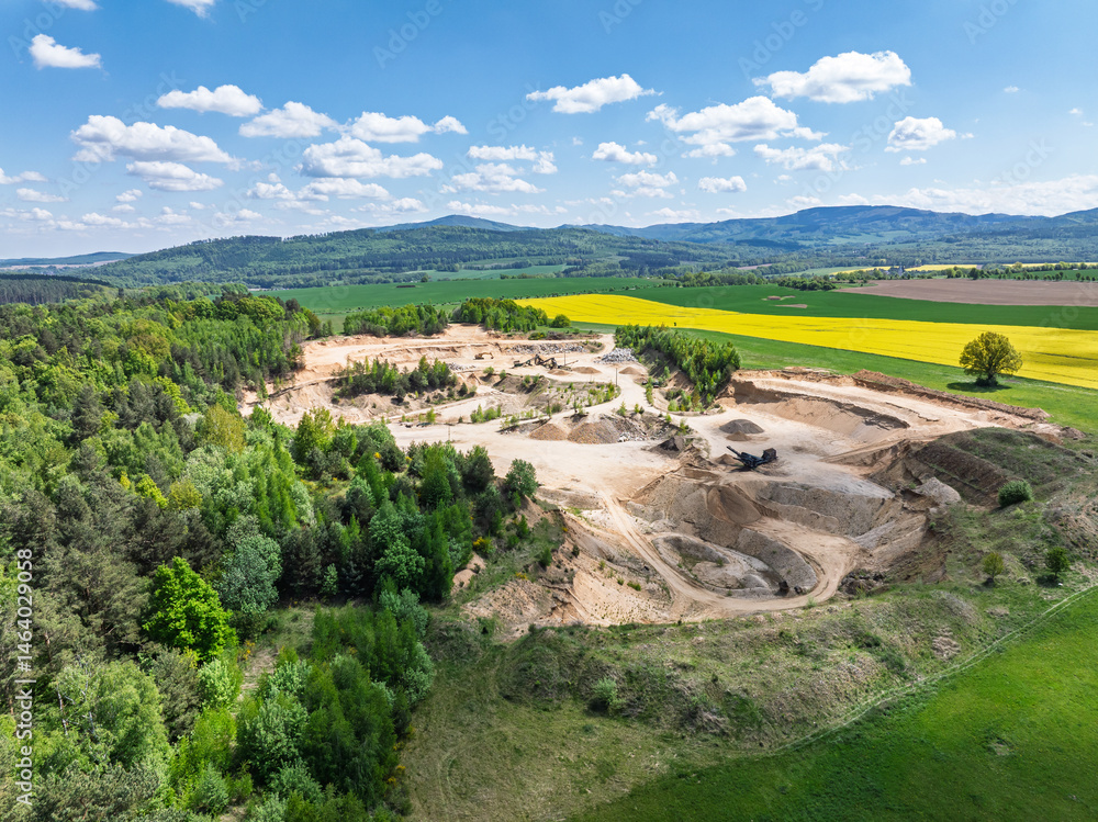 Aerial drone view of sand or gravel quarry surrounded by forest and farmland with distant hills. Aerial Drone view shows an inactive or abandoned quarry embedded in a green landscape.