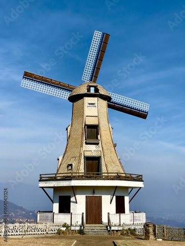 Windmill Under Blue Sky with Mountain View. Traditional windmill on a hilltop with clear blue sky and distant mountains. Bright daytime scene with architectural focus.