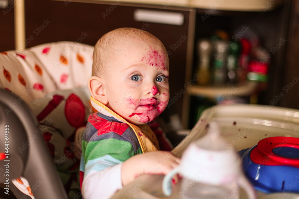Naklejka premium Dirty baby boy while eating boiled beetroot sitting in high chair. Lifestyle photo of parent's daily routine. Introducing complementary foods, first solids.