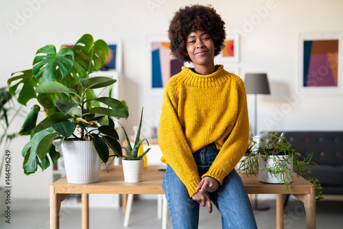 A young woman in a yellow sweater sits on a table surrounded by houseplants. She is smiling in her bright, modern home.