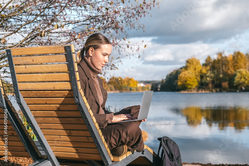 Young woman working remotely by a lake on a laptop. Digital nomad lifestyle, freedom, nature, mindful work, modern remote job. Tranquil eco setting for focus, balance and slow living..