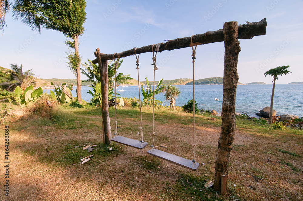 Fototapeta premium A rustic wooden swing set with two swings overlooking a scenic ocean view, surrounded by lush greenery and clear blue sky.