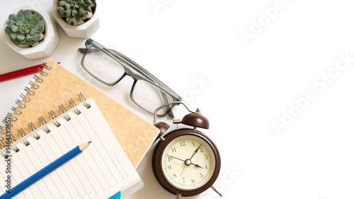 A notebook and pencils sits on a table with an alarm clock and a pair of glasses, isolated on white