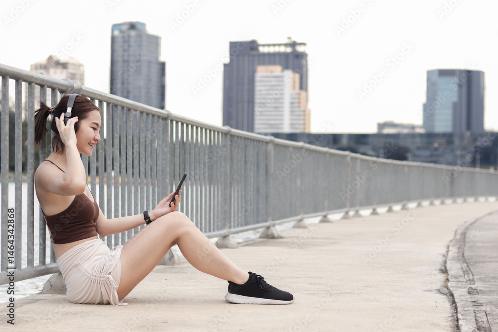 Short haired sporty girl in sportswear woman uses smartphone to listen to music while take a break from exercise outdoor in modern city. Happy beautiful Asian fitness woman working out outside. Short haired sporty girl in sportswear woman uses smartphone to listen to music while take a break from exercise outdoor in modern city. Happy beautiful Asian fitness woman working out outside.