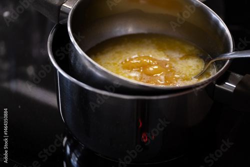 Photos Smooth, fully melted butter in a saucepan over a double boiler setup on a black stovetop, with a spoon partially visible for stirring