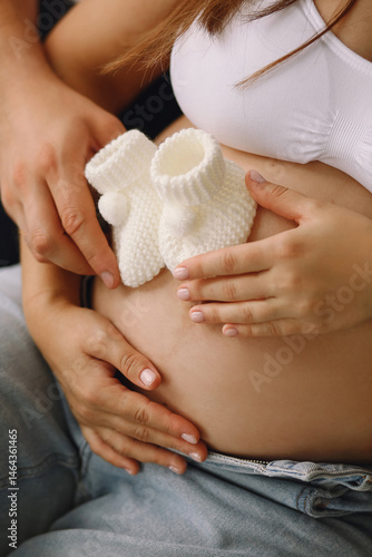 Close up photo of pregnant couple holding white knitted baby socks near pregnant belly indoor. Family concept. The concept of happy parenthood