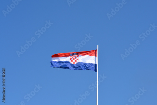 Obraz na płótnie The Croatian national flag, featuring red, white, and blue horizontal stripes and the checkered coat of arms, waves gently against a clear blue sky w salonie