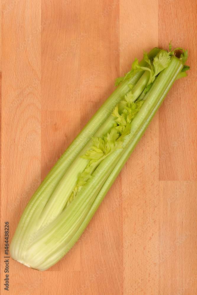 A single stalk of fresh, light green celery with leaves rests diagonally on a wooden cutting board