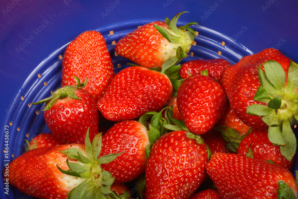 A close-up shows fresh red strawberries with green stems inside a blue colander