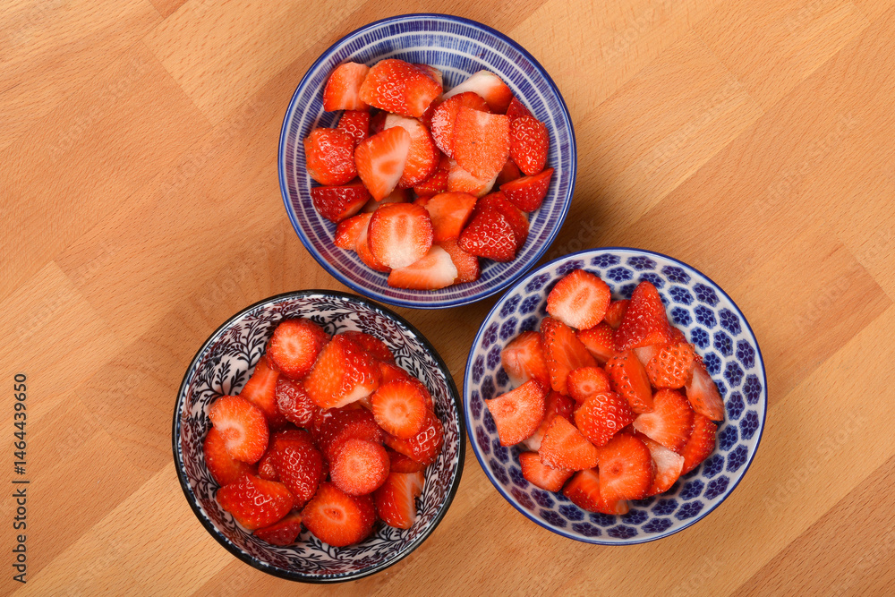 Three small, patterned bowls filled with cut fresh strawberries are arranged in a triangular formation on a light brown wooden surface