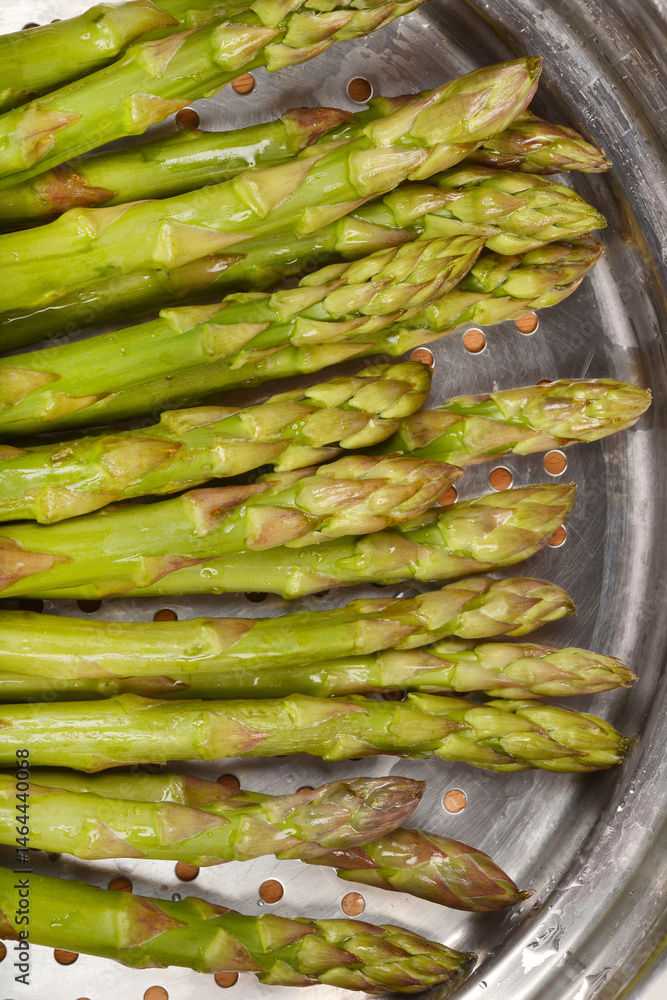 A close-up shows fresh, green asparagus spears arranged inside a metal steamer basket with visible condensation, ready for cooking