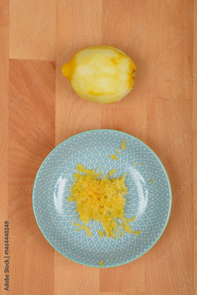 A peeled lemon sits above a light blue decorative plate containing freshly grated yellow lemon zest, all on a light brown wooden surface