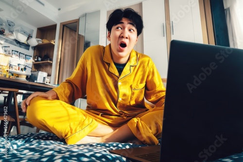 A asian guy wearing yellow pajamas, sitting on a bed, holding his laptop and his expression looks so stressed looking at the laptop.