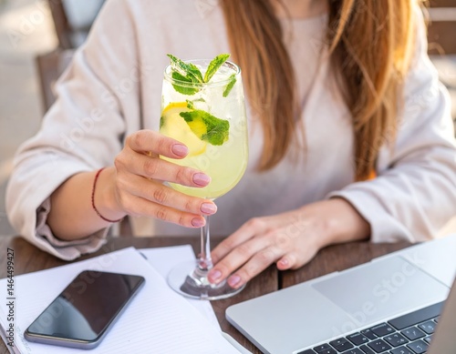 Young woman at outdoor cafe with lemonade, laptop, phone, and notebook, wearing light sweater.
