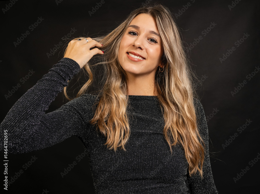 Fototapeta premium Cheerful young woman with long blonde hair playing with her hair while smiling, wearing a sparkly black dress against a dark studio background