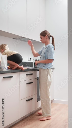 A tired mother is cooking lunch at home in the kitchen, keeping an eye on her restless child climbing onto the kitchen counter. An exhausting routine and daily household chores of everyday family life