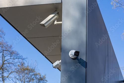 Принти на полотні Security cameras mounted on modern building exterior, capturing surveillance footage, with clear blue sky and tree branches in the background