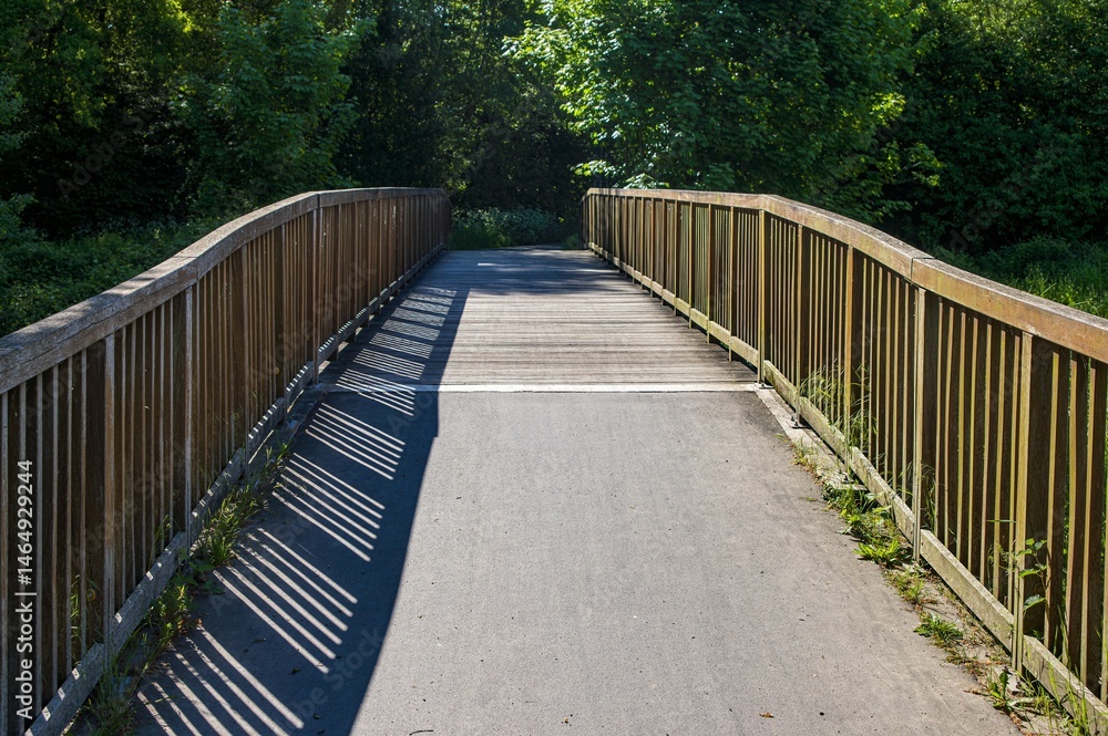 Wooden bridge with shadows stretching across the asphalt path in a park Wooden bridge with shadows stretching across the asphalt path in a park