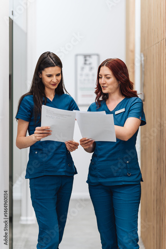 Two female doctors in blue scrubs are standing in a modern hospital corridor, carefully reviewing patient medical records and discussing treatment strategies